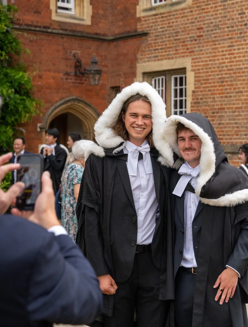 Two graduates smiling together in academic gowns outside a college building