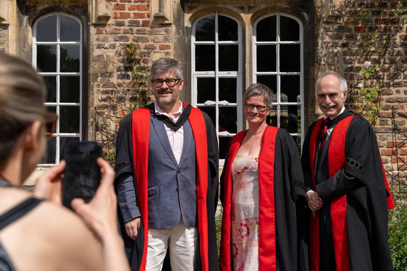 Three people posing in academic dress as a guest takes their photograph