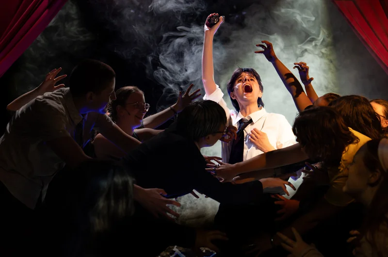 An ensemble reaching towards a performer in a white shirt amid stage smoke and red curtains