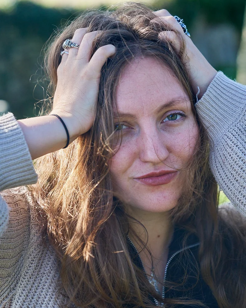 A person looking into the camera outdoors with their hands resting in their hair